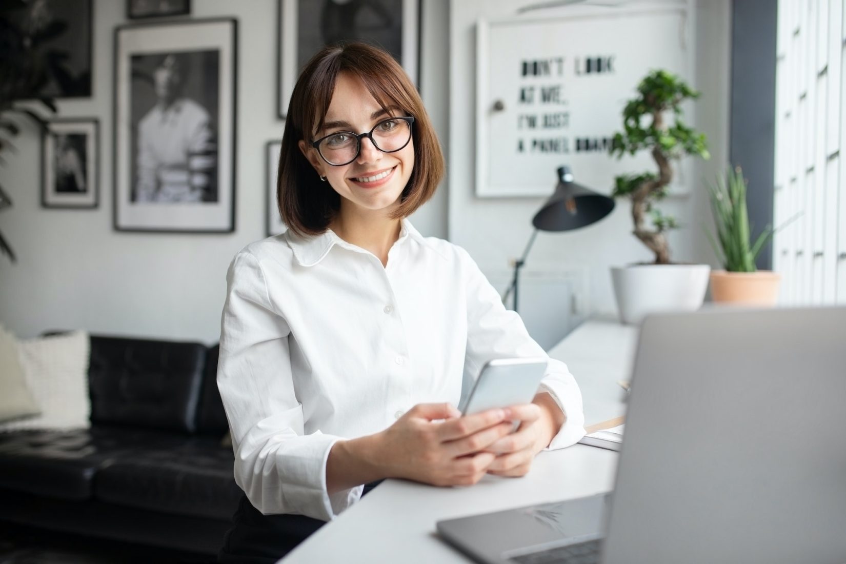 Cheerful smiling freelance lady using cellphone sitting in front of laptop, working in coworking